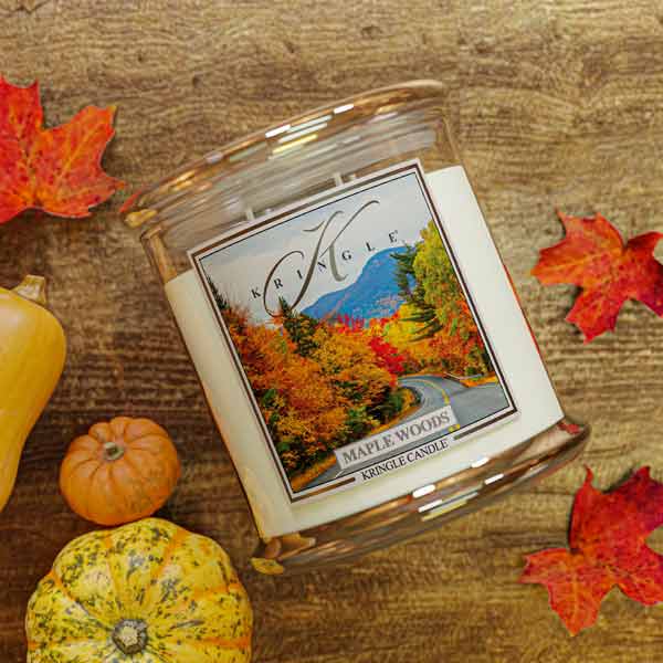 Kringle Candle Maple Woods scented 4-wick candle jar on a wooden surface with autumn leaves and pumpkins