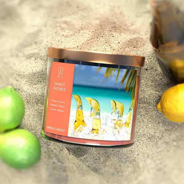 A three-wick candle in a clear glass jar with a bronze lid and a tropical beach-themed label. The label features a serene shoreline with golden sand, turquoise water, and a bright blue sky. The candle’s wax is soft coral, and the wicks are unlit. The scent notes listed are orange, lemon, peach, mandarin, mango, and red currant.
