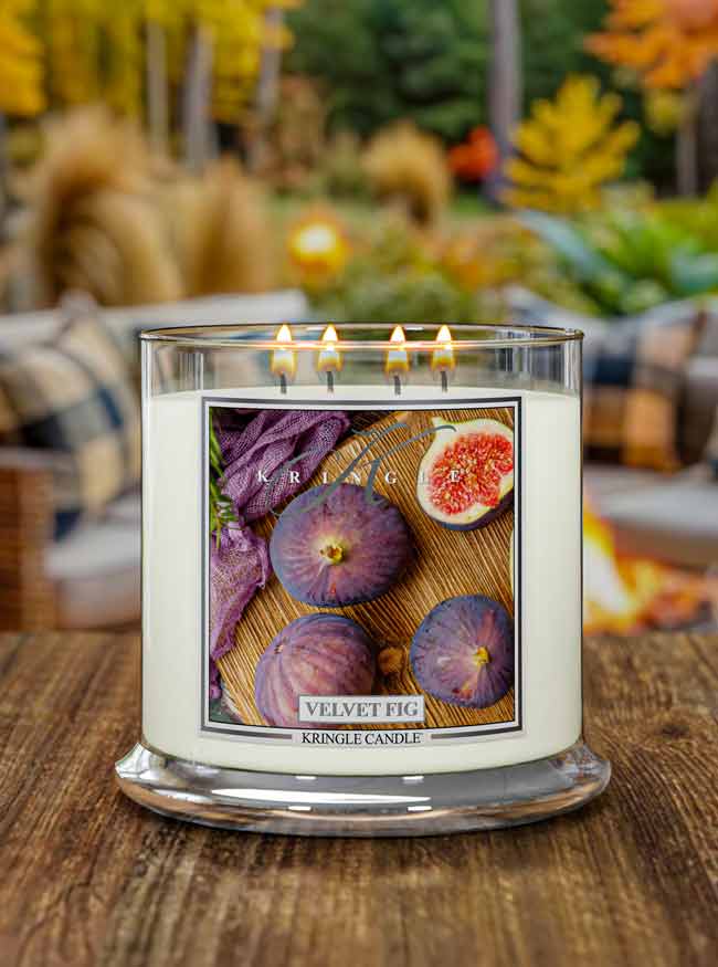 Lit 4-wick candle jar with label showing whole and halved figs on a wooden surface, next to a woven purple cloth.