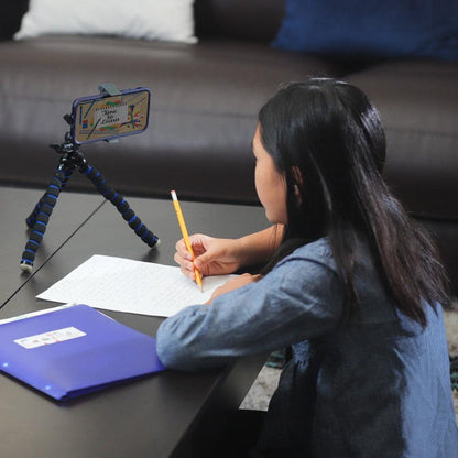 A young girl writes on paper at a low table. In front of her, a smartphone is held by an 11 Adjustable Tripod Mount with Phone Holder, displaying Ultimate Science Class. A blue folder rests beside her on the table.