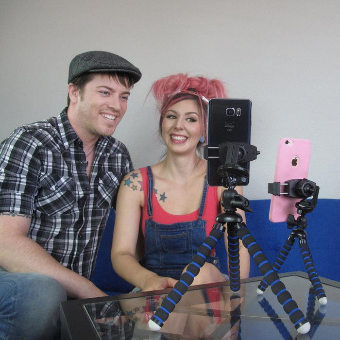 A smiling man and woman sit on a blue couch, recording or livestreaming with two smartphones held by 11 Adjustable Tripod Mounts with Phone Holders atop a glass table.