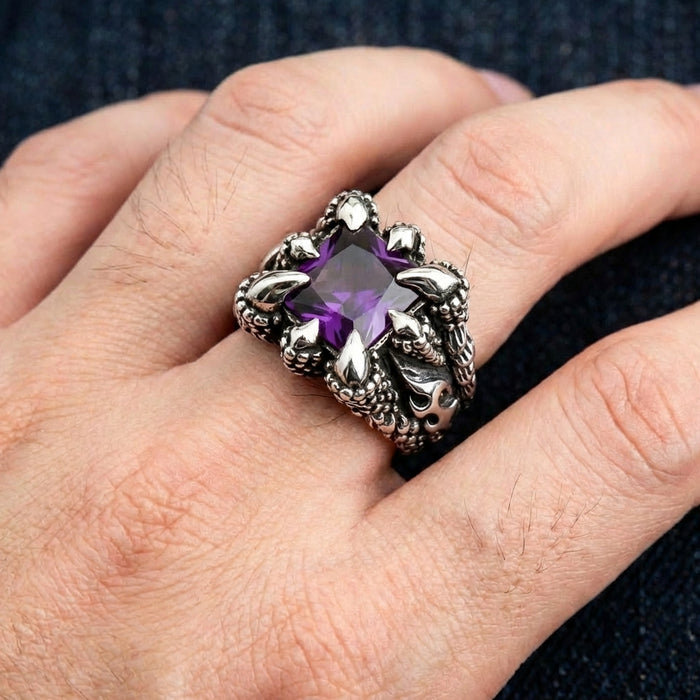 Gothic silver ring with purple amethyst gemstone held by dragon claws worn on a man's finger against dark fabric background