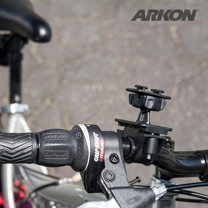 Close-up of a bicycle handlebar with the Bike or Motorcycle Handlebar Mounting Pedestal - Dual-T Compatible attached. Part of the bike seat and blurred pavement are visible in the background, with the Arkon logo in the upper right corner.