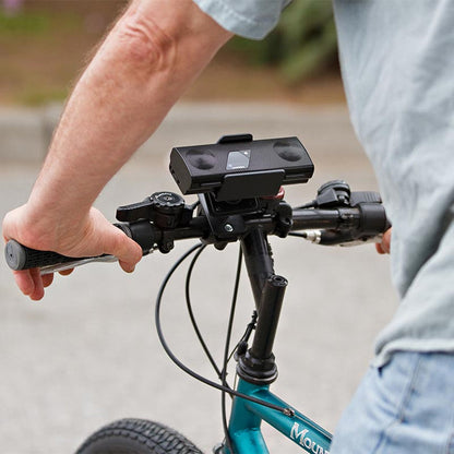 A person rides a bicycle, gripping the handlebars where a black portable speaker is attached using the Bike or Motorcycle Handlebar Mounting Pedestal - Dual-T Compatible, as seen from above and behind.
