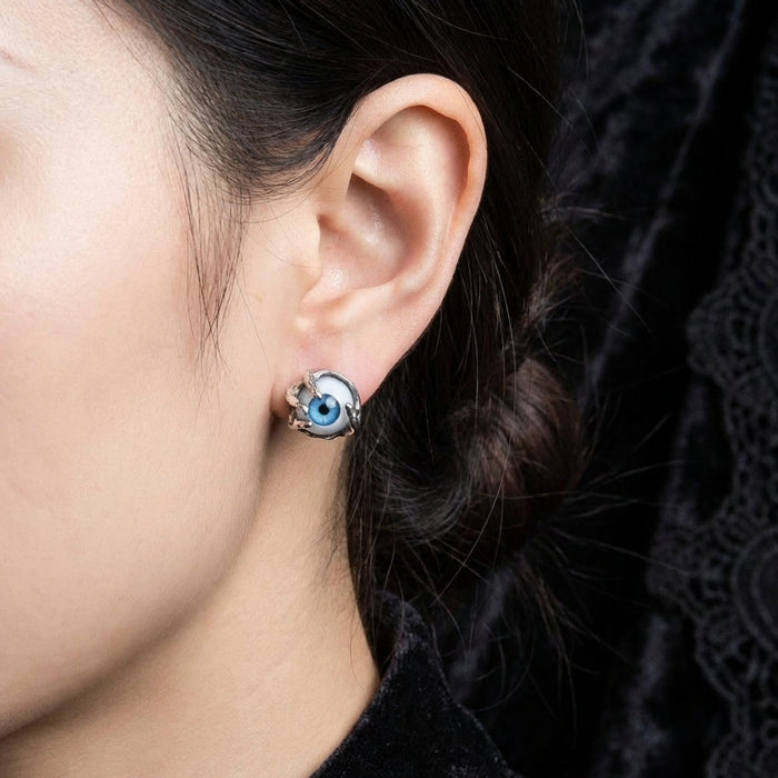 Close-up side profile of a woman wearing a gothic silver stud earring featuring a realistic blue eyeball gripped by metal claws, set against a dark textured background.