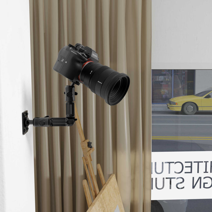 A DSLR camera is attached to the Camera Wall Mount with Multi-Angle Arm for Security Camcorders & Cameras, directed at a flat surface below. Beige curtains and part of a framed picture with a yellow car and STUDIO are behind it.