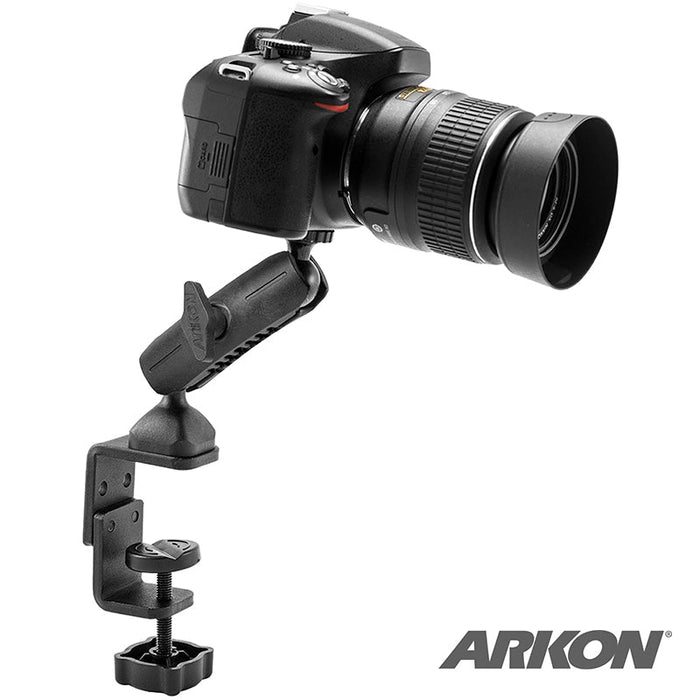 A Clamp Camera Mount for Nikon, Sony, Samsung, Canon, Olympus, and Panasonic Cameras with an adjustable c-clamp is secured to a table edge holding a DSLR. The Arkon logo appears in the bottom right corner.