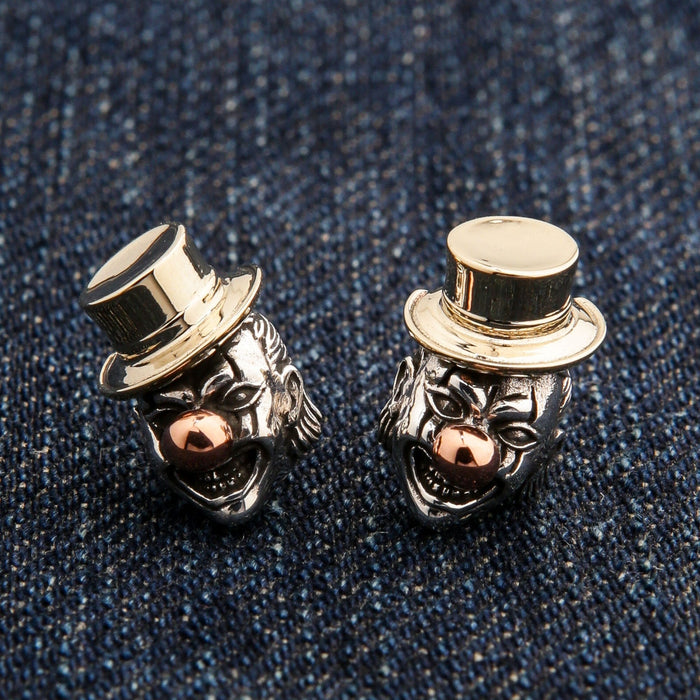 The pair of tri-metal evil clown earrings with brass hats and copper noses on a dark denim surface.