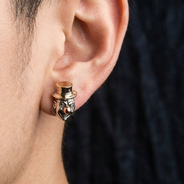 Close-up of a person's ear wearing a detailed silver and gold-tone Joker clown stud earring featuring a creepy face with a golden top hat and nose, set against a dark blurred background.