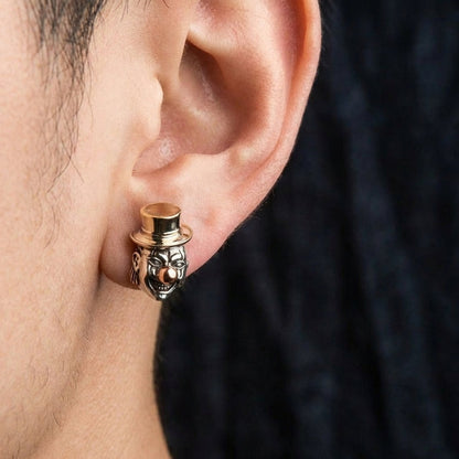 Close-up of a person's ear wearing a detailed silver and gold-tone Joker clown stud earring featuring a creepy face with a golden top hat and nose, set against a dark blurred background.