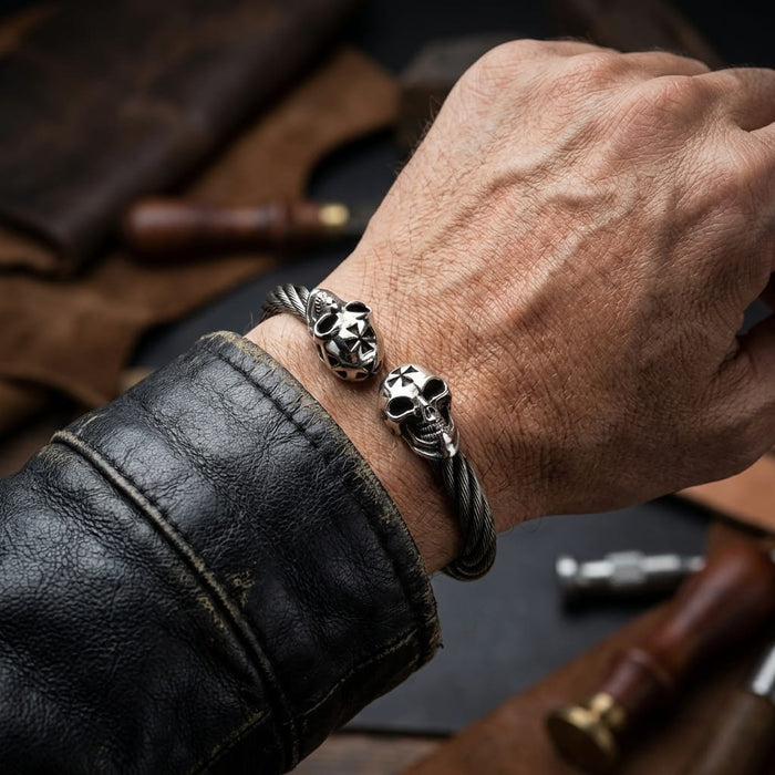 Close-up of a man's wrist wearing a rugged silver twisted cable biker cuff bracelet featuring two skull ends with forehead crosses, styled with a distressed black leather jacket against a workshop background with leather crafting tools.