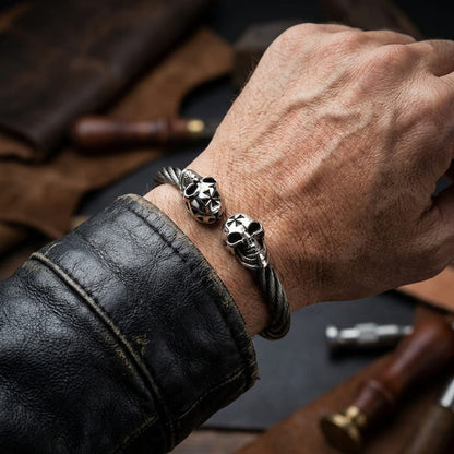 Close-up of a man's wrist wearing a rugged silver twisted cable biker cuff bracelet featuring two skull ends with forehead crosses, styled with a distressed black leather jacket against a workshop background with leather crafting tools.