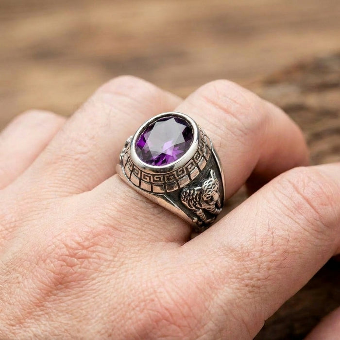 Close-up of a men's sterling silver biker ring worn on a finger, featuring a large oval purple stone, a Greek key pattern around the bezel, and a detailed tiger relief carving on the side shank.