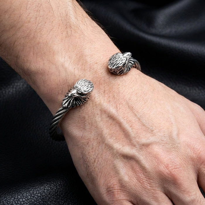 Silver eagle head cuff bracelet with twisted cable band worn on a man's wrist against black leather background, featuring detailed eagle sculptures on both ends in oxidized sterling silver finish