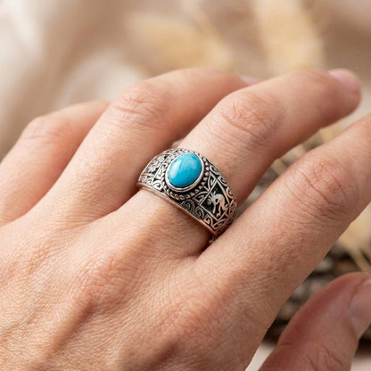Close-up of a person's hand wearing an ethnic oxidized sterling silver ring featuring a vibrant oval turquoise gemstone and intricate Kokopelli engravings, set against a soft, warm blurred background.