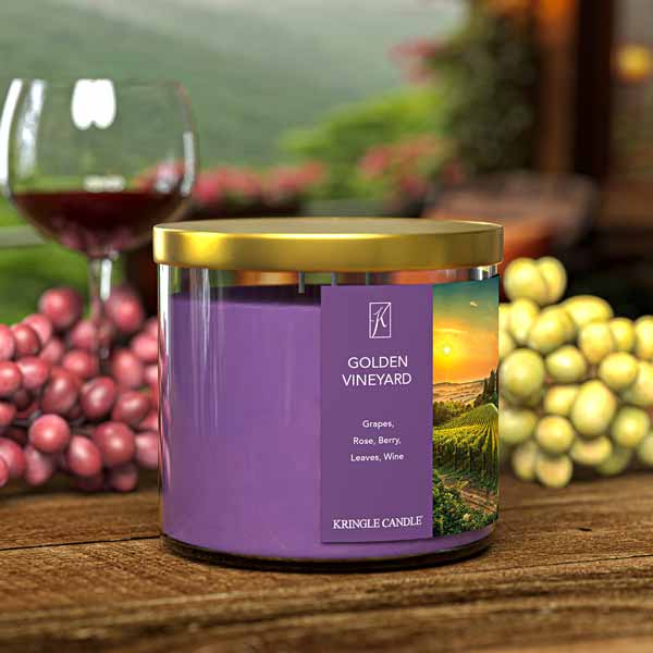 A three-wick candle in a clear glass jar with a gold lid and a scenic vineyard-themed label. The label features a picturesque vineyard at sunset with rolling green hills and a golden sky. The candle’s wax is deep purple, and the wicks are unlit. The scent notes listed are grapes, rose, berry, leaves, and wine.