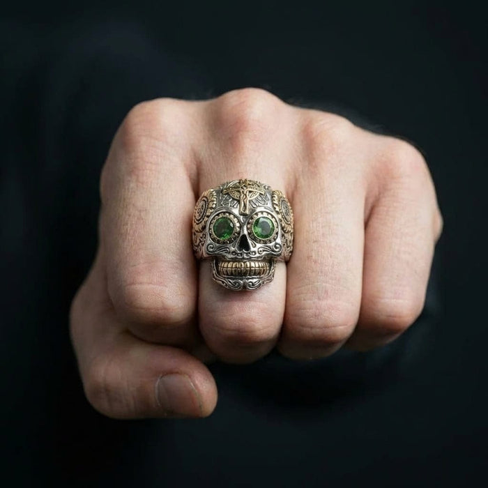 Close-up of a men's sterling silver sugar skull ring featuring green gemstone eyes and gold accents on the forehead cross and teeth, worn on a clenched fist against a dark background.