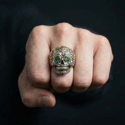 Close-up of a men's sterling silver sugar skull ring featuring green gemstone eyes and gold accents on the forehead cross and teeth, worn on a clenched fist against a dark background.