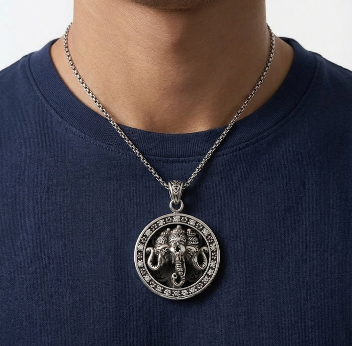 Oxidized silver Ganesha pendant necklace with round medallion design featuring intricate elephant god relief and floral border, worn by a man on a silver chain over a navy blue t-shirt