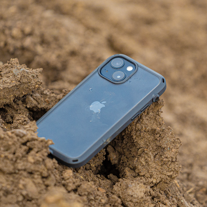 A Catalyst Case US iPhone 14 - Influence Case with a blue finish and visible Apple logo lies face down on rough brown dirt, partially buried. Dual camera lenses and rugged design hint at protection for outdoor adventures.