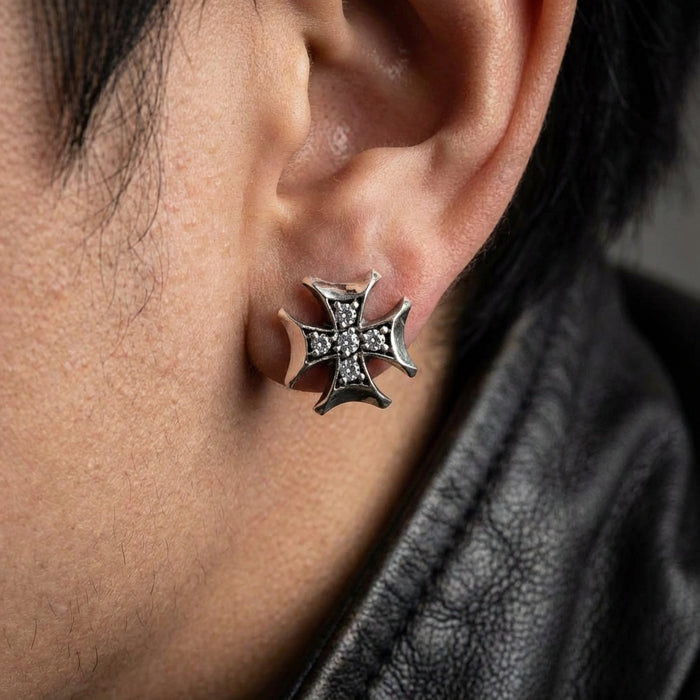 Close-up editorial shot of a rocker wearing 12mm iced out iron cross earrings under soft studio lighting.