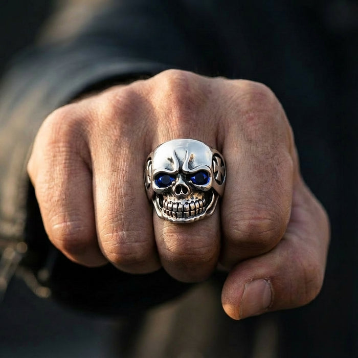 Close-up of a man's clenched fist wearing a heavy sterling silver skull ring featuring deep blue gemstone eyes and an Iron Cross emblem on the side.