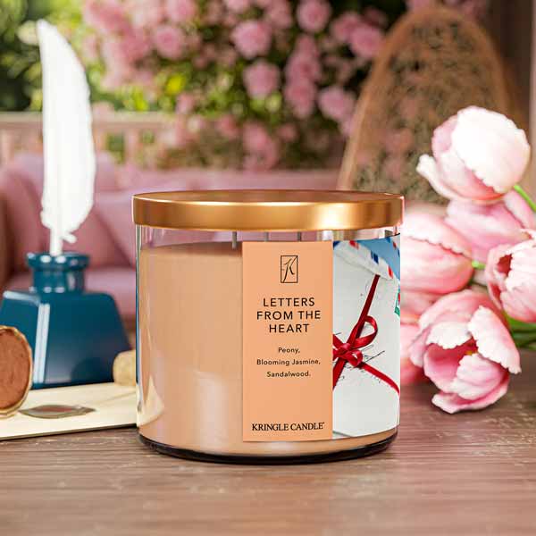 A three-wick candle in a clear glass jar with a bronze lid and a sentimental-themed label. The label features a bundle of vintage love letters tied with a red ribbon, with USA heart stamps on top. The candle’s wax is a soft peach shade, and the wicks are unlit. The scent notes listed are peony, blooming jasmine, and sandalwood.