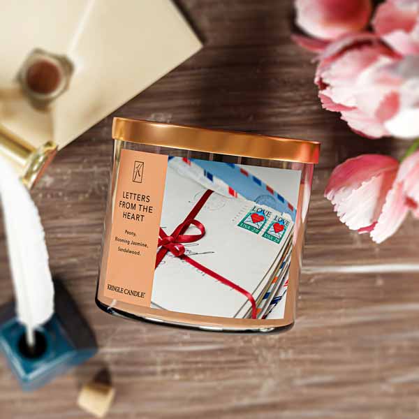 A three-wick candle in a clear glass jar with a bronze lid and a sentimental-themed label. The label features a bundle of vintage love letters tied with a red ribbon, with USA heart stamps on top. The candle’s wax is a soft peach shade, and the wicks are unlit. The scent notes listed are peony, blooming jasmine, and sandalwood.
