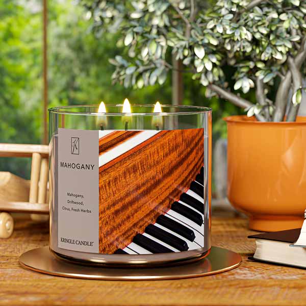 A three-wick lit candle in a clear glass jar with a luxurious wood and music-themed label. The candle’s wax is a rich brown, and all three wicks are burning.