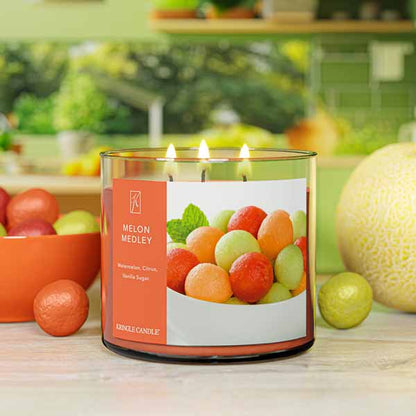 A three-wick lit candle in a clear glass jar with a fresh fruit-themed label. The candle’s wax is warm orange, and all three wicks are burning.