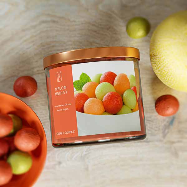 A three-wick candle in a clear glass jar with a bronze lid and a fresh fruit-themed label. The label features a bowl filled with colorful melon balls, including watermelon, cantaloupe, and honeydew. The candle’s wax is warm orange, and the wicks are unlit. The scent notes listed are watermelon, citrus, and vanilla sugar.