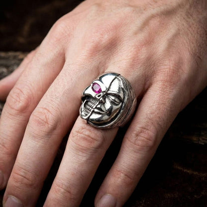Close-up of a male hand wearing a heavy silver Memento Mori two-face ring, featuring a split design with a half-skull sporting a pink gemstone eye and a half-serene human face, set against a dark textured background.