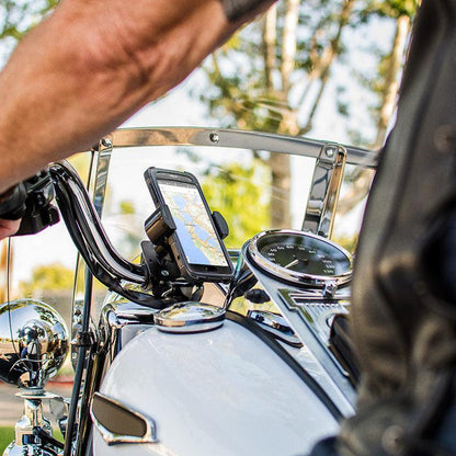 Close-up of a person’s arm on the handlebar of a motorcycle equipped with the RoadVise® Holder with Handlebar Mount displaying a map on their phone. The speedometer, chrome details, and blurred trees are visible in the background.