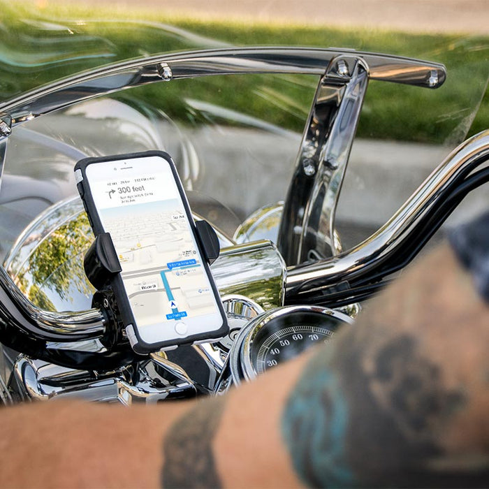 A smartphone with a GPS map is secured on motorcycle handlebars using the RoadVise® Motorcycle Phone Mount - Black Aluminum. A tattooed arm appears in the foreground, while chrome parts of the motorcycle reflect the light.