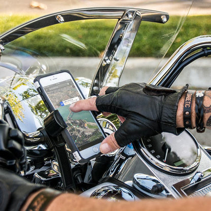 A person wearing black leather gloves uses a GPS app on a smartphone secured in a RoadVise® Motorcycle Phone Mount with Strap attached to the handlebars of a motorcycle, with the dashboard and chrome details visible.
