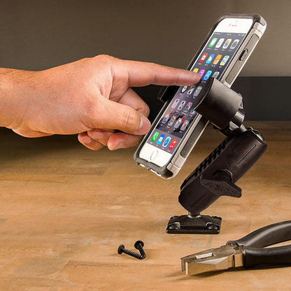 A hand touches a smartphone on a RoadVise® Phone Holder with Drill Base and Adjustable Arm attached to a table, with two loose screws and pliers lying nearby on the wooden surface.