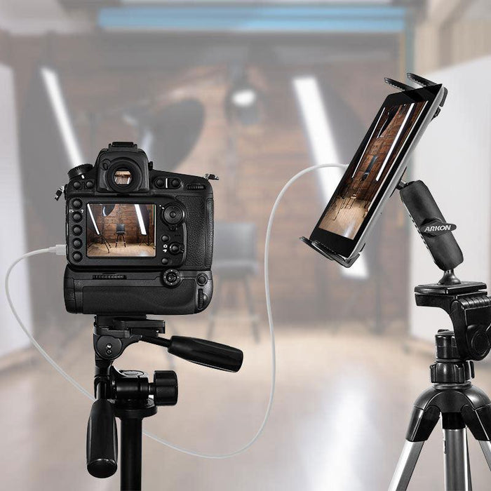A professional camera and a tablet mounted on separate tripods, with the tablet using the Slim-Grip® Tripod Tablet Mount with 3.75 Shaft, are connected by a cable in a studio with blurred lights and equipment in the background.