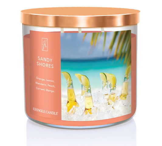 A three-wick candle in a clear glass jar with a bronze lid and a tropical beach-themed label. The label features a serene shoreline with golden sand, turquoise water, and a bright blue sky. The candle’s wax is soft coral, and the wicks are unlit. The scent notes listed are orange, lemon, peach, mandarin, mango, and red currant.