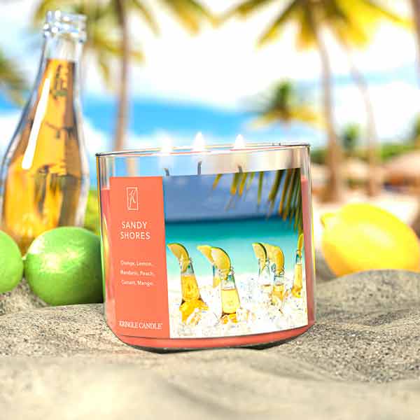 A three-wick lit candle in a clear glass jar with a tropical beach-themed label. The candle’s wax is soft coral, and all three wicks are burning.