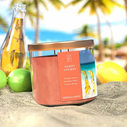 A three-wick candle in a clear glass jar with a bronze lid and a tropical beach-themed label. The label features a serene shoreline with golden sand, turquoise water, and a bright blue sky. The candle’s wax is soft coral, and the wicks are unlit. The scent notes listed are orange, lemon, peach, mandarin, mango, and red currant.