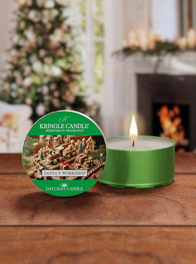 Green Kringle Candle with Santa's Workshop label on a wooden surface, Christmas tree and fireplace in the background.