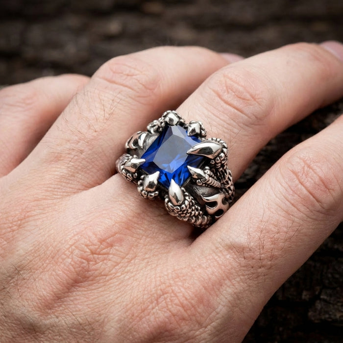 Gothic silver claw ring featuring a large rectangular blue sapphire gemstone gripped by dragon talons, worn on a man's finger with dark textured background