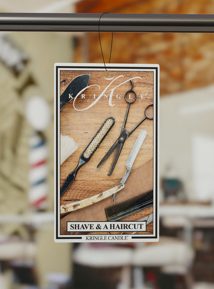 Kringle Candle air freshener with barber tools on a wooden surface, blurred barbershop background