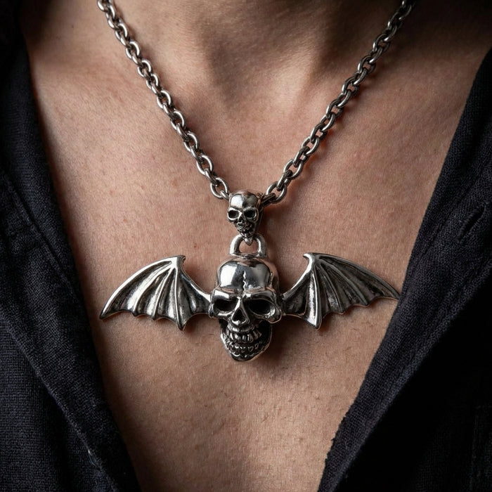 Close-up of a person wearing a heavy silver gothic necklace featuring a detailed skull pendant with bat wings, attached to a smaller skull bail on a thick chain, styled with an open black shirt.
