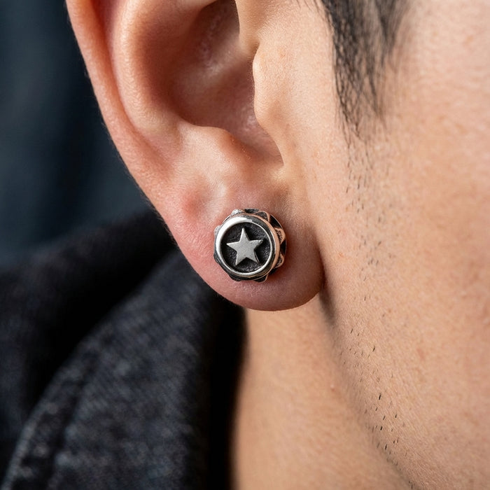 Close-up of a man wearing an oxidized silver star stud earring with a raised five-point star design set in a round black background, styled with casual denim clothing