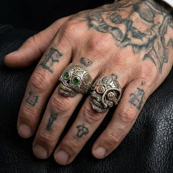 Two men's sterling silver steampunk skull rings worn on a tattooed hand, featuring mechanical gear details, green gemstone eyes, and gothic biker jewelry design against a black leather background.