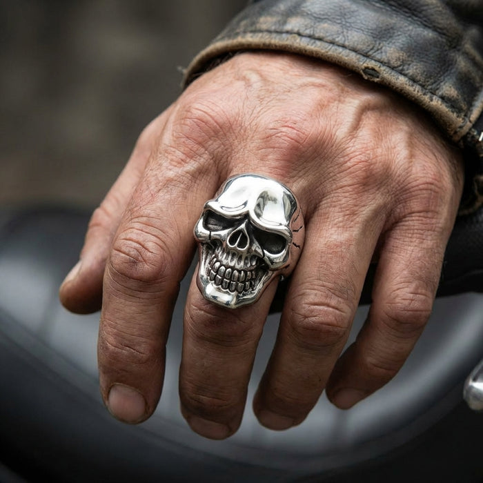 Large silver skull ring with detailed teeth and hollow eyes worn on a man's finger with weathered leather jacket sleeve, showcasing rugged biker jewelry style against a motorcycle background