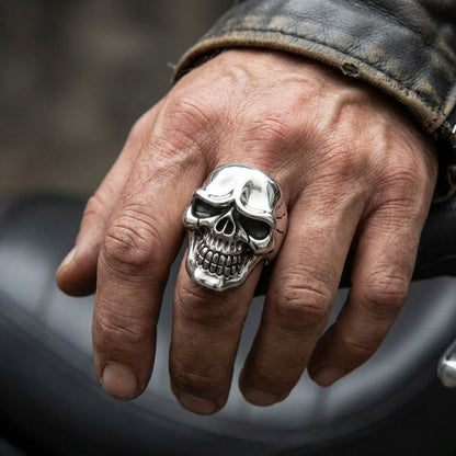 Large silver skull ring with detailed teeth and hollow eyes worn on a man's finger with weathered leather jacket sleeve, showcasing rugged biker jewelry style against a motorcycle background