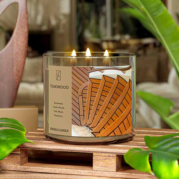 A three-wick lit candle in a clear glass jar with a sophisticated wood-themed label. The candle’s wax is rich brown, and all three wicks are burning.