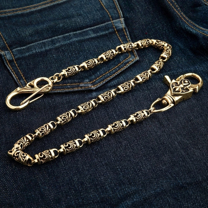 Tribal scrollwork brass chain laid open on denim, snap hook at one end and the carved lobster claw at the other.